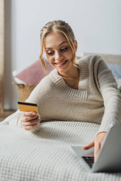 potěšená žena ve svetru držící kreditní kartu a pomocí notebooku při online nakupování v ložnici  - Fotografie, Obrázek