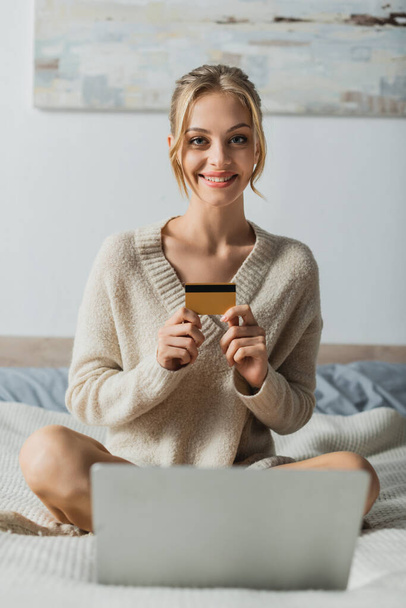 cheerful woman holding credit card near laptop while doing online shopping in bedroom  - Photo, image