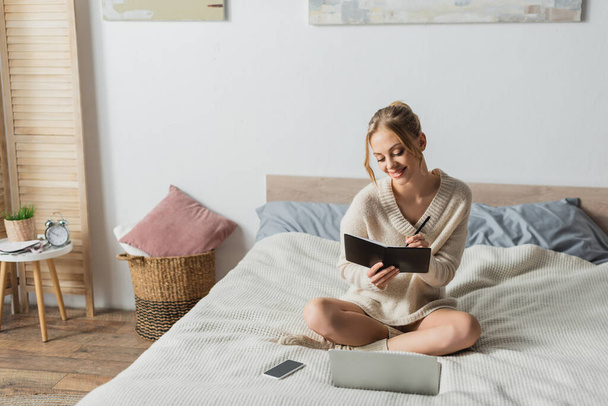 cheerful blonde woman taking notes near gadgets on bed in modern apartment  - Fotó, kép