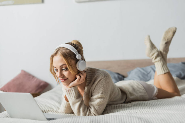 fiatal szőke nő vezeték nélküli fejhallgató néz filmet laptop a hálószobában  - Fotó, kép