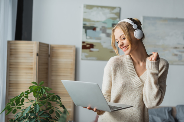 joyful young woman in wireless headphones listening music and holding laptop in modern bedroom  - Foto, imagen