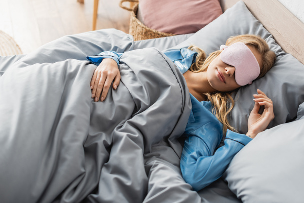 από πάνω όψη ξανθιάς γυναίκας με ροζ μάσκα ύπνου και μπλε πιτζάμα αναπαύεται στο κρεβάτι  - Φωτογραφία, εικόνα