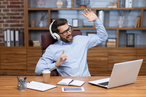 Mature boss investor at workplace inside home office dancing having fun listening to music in headphones, businessman in shirt resting and happy sitting at table using laptop at work. - Photo, Image
