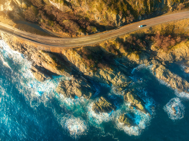 Вид с воздуха на дорогу, скалистое побережье с волнами и камнями на закате в Лофотенских островах, Норвегия. Пейзаж с красивой дорогой, прозрачная голубая вода, камни. Вид сверху с дрона шоссе летом - Фото, изображение