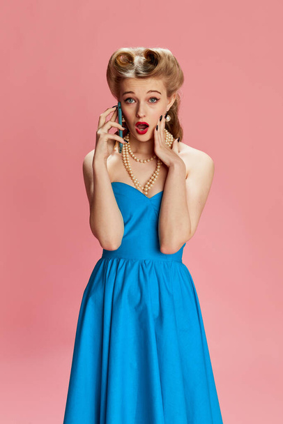 Portrait of beautiful young girl in blue dress and stylish hairstyle emotionally talking on phone against pink studio background. Concept of retro fashion, beauty, 50s, 60s. Pin-up style - 写真・画像