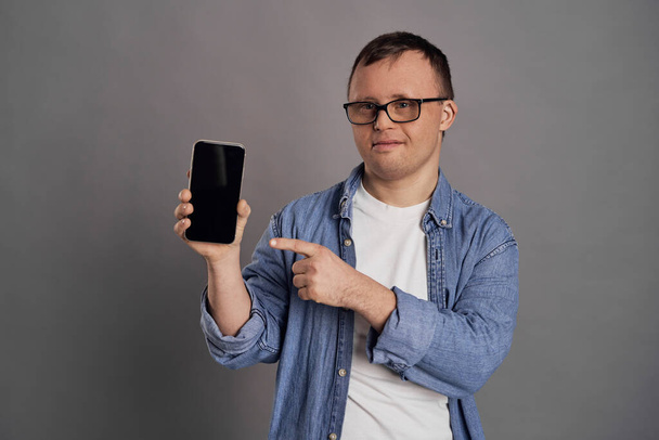 Adult man with down syndrome showing empty screen of mobile phone on gray background - Фото, изображение