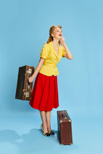 Χαρούμενος ταξιδιώτης. Πορτρέτο της όμορφης νεαρής κοπέλας με κίτρινη μπλούζα και κόκκινη φούστα που ποζάρει με βαλίτσες σε φόντο μπλε στούντιο. Ρετρό μόδα, ομορφιά, ταξίδια, 50, 60 έννοια. Στυλ καρφιτσών - Φωτογραφία, εικόνα