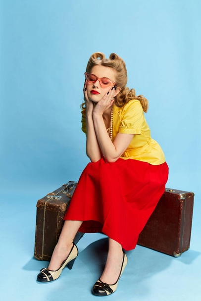 Portrait of beautiful young girl in yellow blouse and red skirt sitting on vintage suitcases against blue studio background. Concept of retro fashion, beauty, travelling, 50s, 60s. Pin-up style - Fotografie, Obrázek