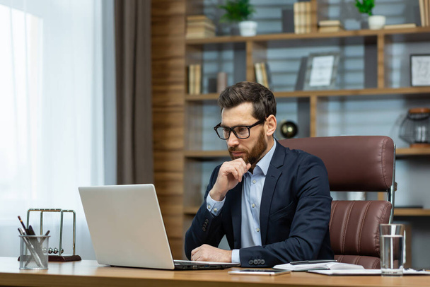 Thinking businessman working sitting at desk, mature adult boss in business suit and beard looking at laptop screen thinking about financial investment decisions inside office. - Фото, зображення