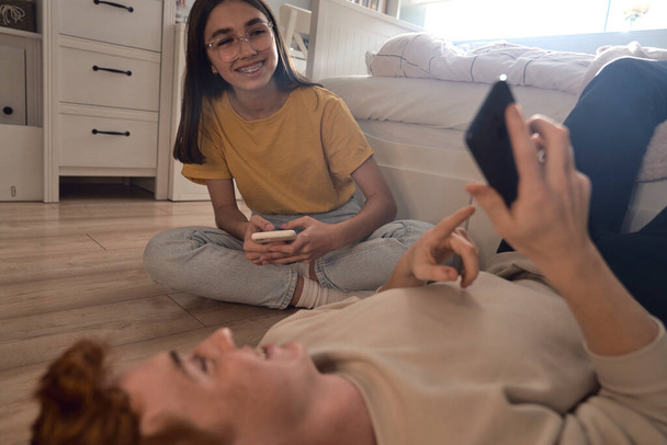 Dos amigos adolescentes caucásicos se divierten mientras usan teléfonos móviles en el dormitorio - Foto, imagen