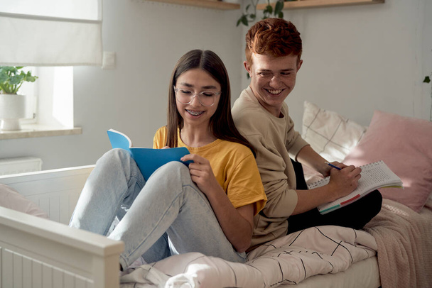 Two caucasian teenagers sitting bed back to back and learning from books with fun  - Photo, Image