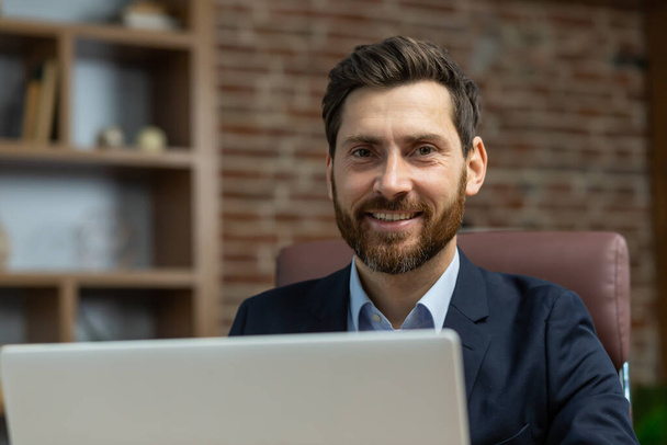 Close up portrait of successful mature adult businessman inside home office, bearded man at work with laptop smiling and looking at camera happy with remote work. - 写真・画像