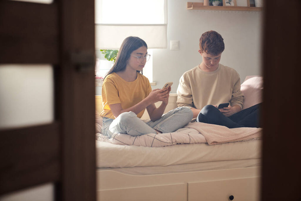 Two caucasian teenage friends sitting on bed in bedroom and browsing phones in silence - Photo, Image