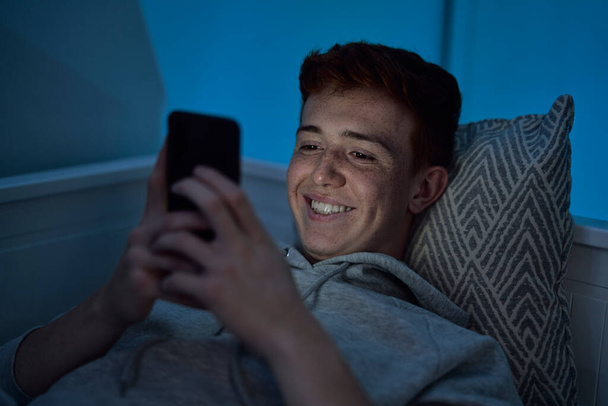 Vidám, kaukázusi tinédzser fiú mobiltelefont használ, miközben éjjel az ágyban fekszik a szobájában.  - Fotó, kép