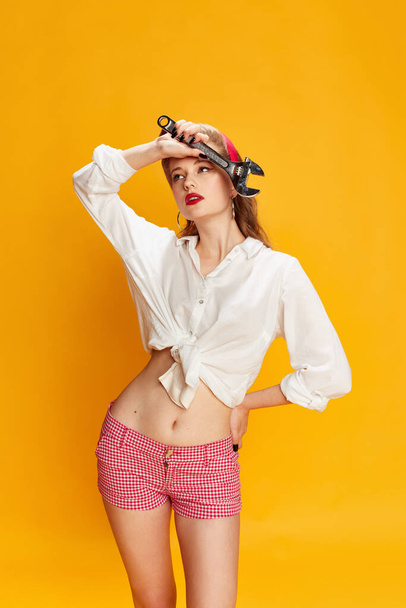 Feeling tired. Portrait of beautiful young girl in white shirt and pink shorts posing with tool, wrench against yellow studio background. Concept of retro fashion, beauty, 50s, 60s. Pin-up style - Zdjęcie, obraz