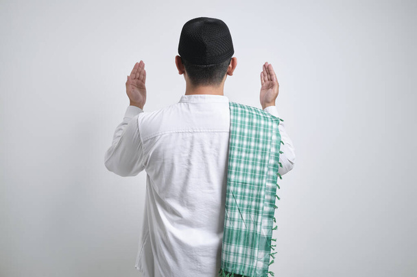 Junger asiatischer muslimischer Mann hebt die Hand und betet rückwärts stehend isoliert vor weißem Hintergrund - Foto, Bild