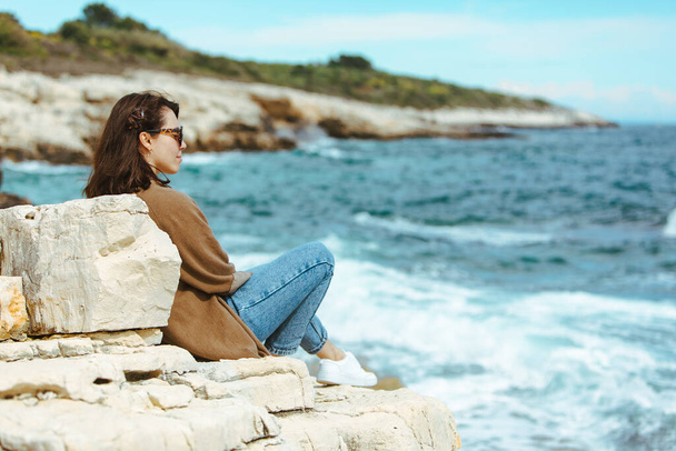 donna seduta sulla scogliera godendo della vista sul mare. Tempo ventoso. Giornata di sole. vacanze estive. copia spazio
 - Foto, immagini