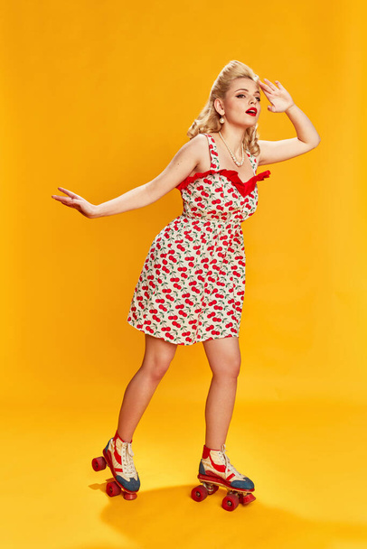 Seeking out. Portrait of beautiful young girl with stylish hairstyle in dress, on rollers posing against yellow studio background. Concept of retro fashion, beauty, 50s, 60s. Pin-up style - Фото, изображение