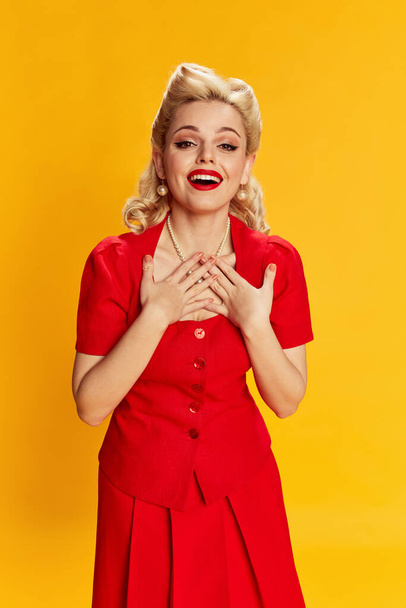 Happy, smiling. Portrait of beautiful young girl with stylish hairstyle in red dress posing against yellow studio background. Concept of retro fashion, beauty, 50s, 60s. Pin-up style - Фото, изображение