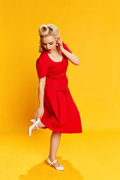 Portrait of beautiful young girl with stylish hairstyle in red suit posing against yellow studio background. Concept of retro fashion, beauty, 50s, 60s. Pin-up style - Photo, image