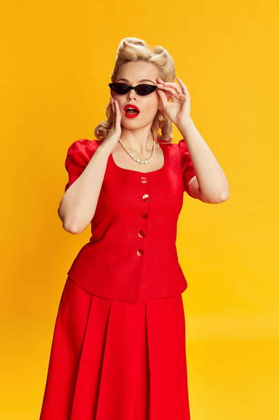 Beautiful young girl with stylish hairstyle in red suit and trendy sunglasses posing with shocked face against yellow studio background. Concept of retro fashion, beauty, 50s, 60s. Pin-up style - Fotoğraf, Görsel