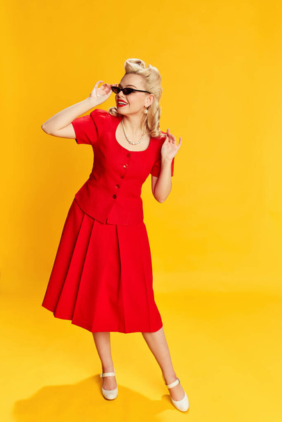 Portrait of beautiful young blonde woman with stylish hairstyle in red suit and trendy sunglasses posing against yellow studio background. Concept of retro fashion, beauty, 50s, 60s. Pin-up style - Fotoğraf, Görsel