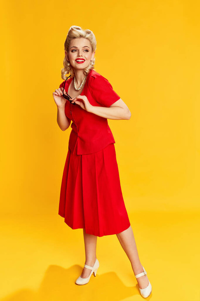 Happy, positive mood. Portrait of beautiful young girl with stylish hairstyle in red suit posing against yellow studio background. Concept of retro fashion, beauty, 50s, 60s. Pin-up style - Фото, изображение