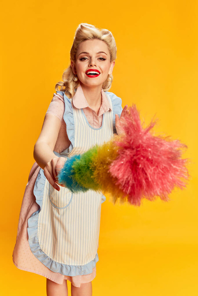 Cleaning. Portrait of beautiful young blonde woman with stylish hairstyle posing with pipidastre against yellow studio background. Concept of retro fashion, beauty, 50s, 60s. Pin-up style - 写真・画像