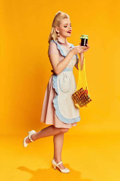 Food shopping. Portrait of beautiful young girl with stylish hairstyle posing against yellow studio background. Concept of retro fashion, beauty, 50s, 60s. Pin-up style - Foto, Imagen