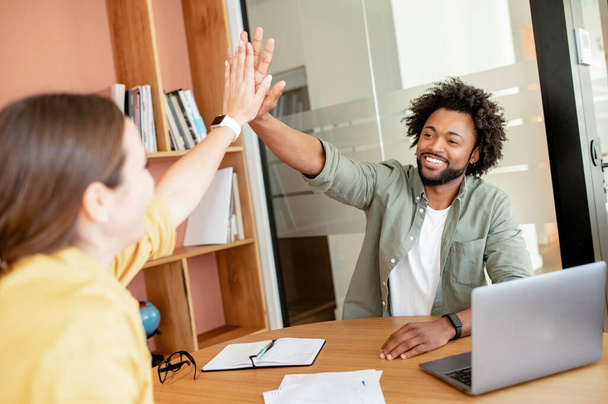 Two diverse employees in smart casual wear giving high five to each other indoors in the contemporary meeting room, celebrating successful collaboration, colleagues support each other - Photo, image
