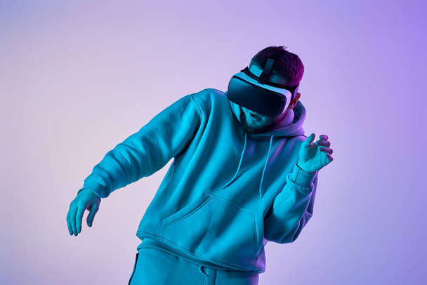 man in sweatshirt using virtual reality glasses and playing video games on blue background. Neon lighting - Photo, Image