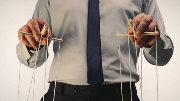 A businessman in a gray shirt and black tie controls a puppet with a wooden manipulator and strings. The puppeteer manipulates the puppet by pulling the ropes with both hands on a white background - Φωτογραφία, εικόνα