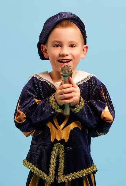 Счастливый мальчик дошкольного возраста в костюме средневекового мальчика, маленький принц поет за микрофоном на светло-голубом фоне. Концепция детских эмоций, сравнение эпох, мода, музыка, искусство, фестиваль - Фото, изображение