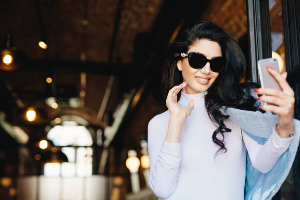 Mulher glamour sorridente em óculos de sol, blusa branca e jaqueta posando na câmera de seu smartphone enquanto faz selfie fotografando a si mesma. modelo elegante feliz posando no café usando telefone celular
 - Foto, Imagem