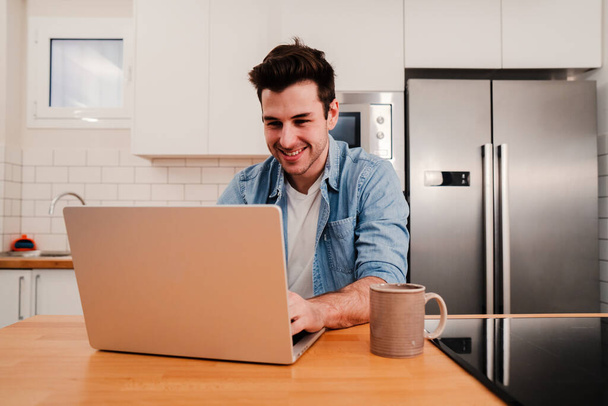 Handsome caucasian man working with a laptop computer at home kitchen drinking coffee and enjoying the morning. Happy young entrepreneur or student searching information on internet. Freelance work - Photo, image