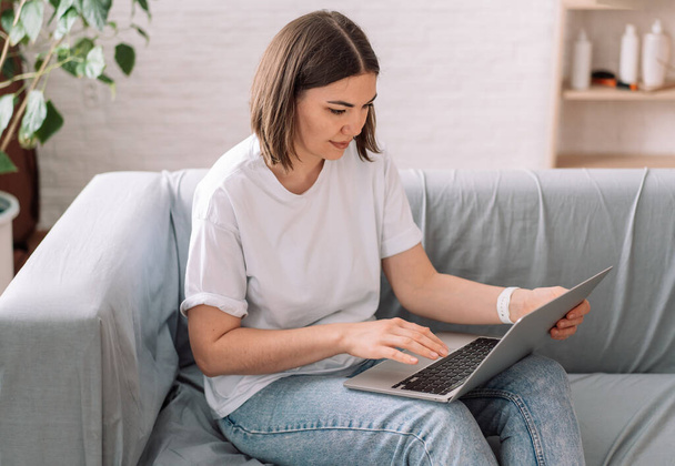 Kobieta siedząca na szarej sofie bawiąca się laptopem. - Zdjęcie, obraz