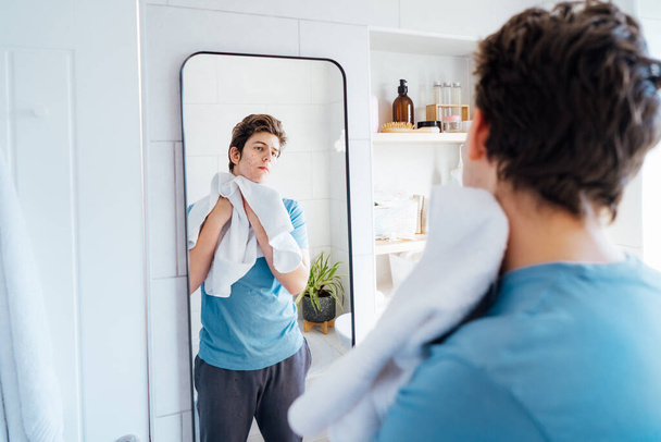 Concentrated caucasian teenage boy with acne problem takes care of his face skin at home. He looking in mirror and wipes face with towel in bathroom. Teenager skin care every day treatment process - Фото, изображение