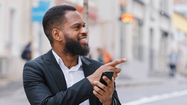 Афро-американский бородатый человек с приложением GPS навигации смартфона потерял в городе ищет адрес с онлайн-приложением карты в мобильном телефоне. Этнический бизнесмен показывает направление пальцем - Фото, изображение