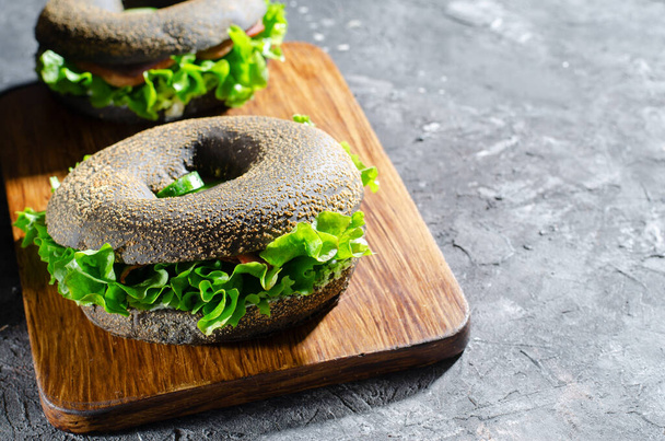 Black Bagels Sandwiches with Cucumbers, Salad and Ham on Wooden Board and Dark Background, Tasty Snack or Breakfast - Photo, Image