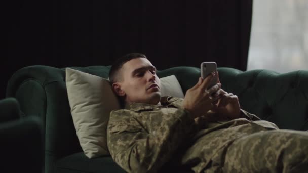 Un joven con uniforme militar yace en el sofá y desplaza la alimentación de su smartphone. Un joven soldado está mirando las noticias en su teléfono y está esperando la llegada de un psicoterapeuta. - Metraje, vídeo
