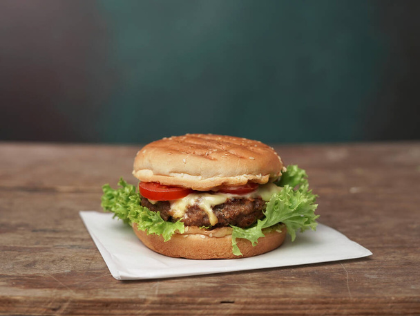 Big Burger βρίσκεται στην τέχνη λευκό χαρτί έναντι ξύλινο τραπέζι. Μια πράσινη σαλάτα και μια κόκκινη ντομάτα κείτονται κοντά στο Burger. - Φωτογραφία, εικόνα