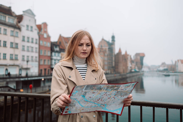 Attraktive junge Touristinnen erkunden die neue Stadt. Blondes Mädchen mit einer Landkarte aus Papier auf der Straße in Danzig. Europa im Herbst bereisen.  - Foto, Bild