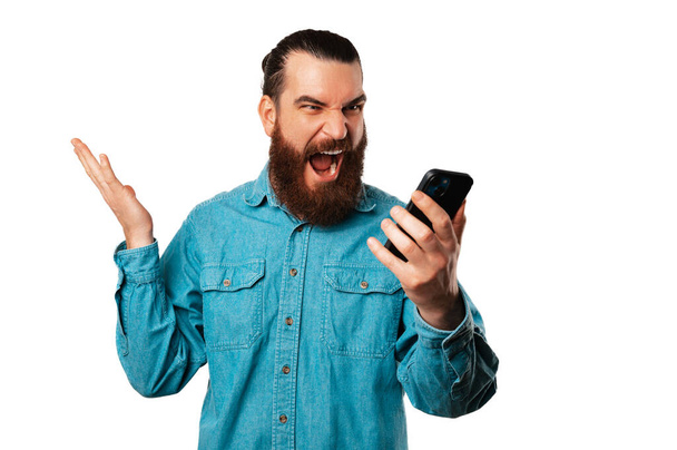 Расстроенный бородатый мужчина кричит на телефон, который он держит во время звонка на белом фоне. - Фото, изображение