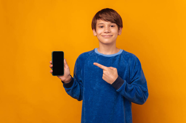 Cheerful teen boy is pointing at the screen of phone he is holding aside over yellow background. - Photo, Image