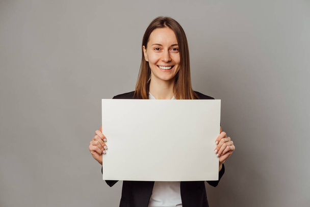 Cheerful young woman wearing jacket holds white paper board with copy space over grey background. - Foto, Imagem