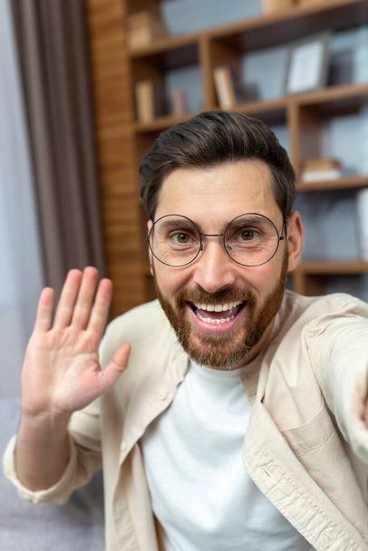 A young man in glasses talks at home on a video call. He holds the phone in his hands, waves at the camera, smiles, says hello. Vertical photo. - Foto, Bild