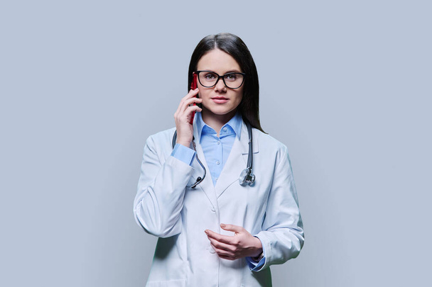Young female doctor talking on mobile phone, on light gray background of studio. Friendly physician talking to patient. Medical staff occupation, technologies mobile applications, health care medicine - Foto, Imagem