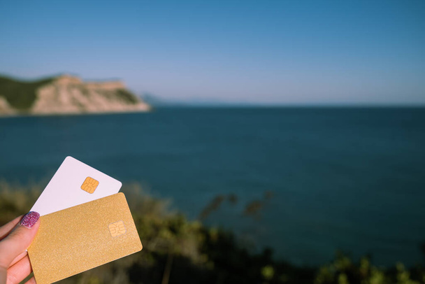 Golden and White Bank Card In Woman Hand On Background Of Scenic View From Arkoudilas Viewpoint, Mountains, Ionian Sea Corfu, Greece. The Concept Of Payment For Relax, Unlimited Possibilities. High - Foto, imagen
