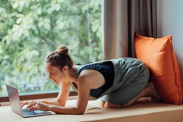 Μια φοιτήτρια ξαπλωμένη στο παράθυρο και δουλεύει στο λάπτοπ. Νεαρή γυναίκα τουρίστρια ξαπλωμένη στο περβάζι του παραθύρου και χρησιμοποιώντας υπολογιστή στο δωμάτιο ξενοδοχείου ή πανεπιστημιούπολης. - Φωτογραφία, εικόνα