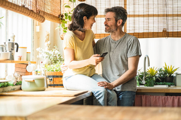 casal maduro, mulher sentada no balcão da cozinha, olhando para o telefone inteligente feliz sorrindo - Foto, Imagem